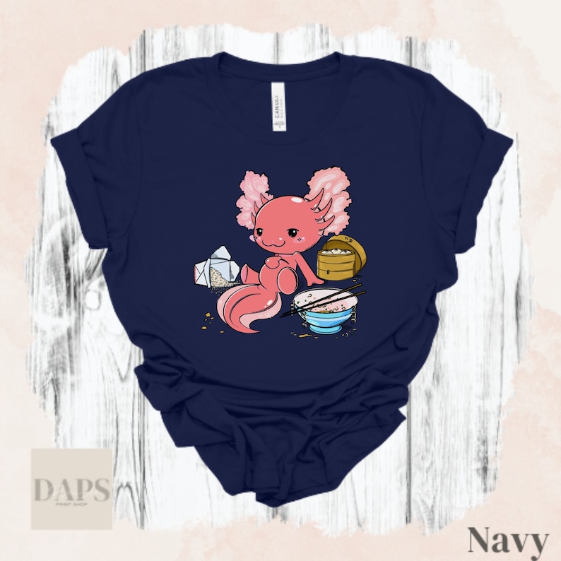 Peut inclure: Un t-shirt bleu marine avec une illustration de dessin anim&eacute; d'un axolotl portant un chapeau rose et tenant des baguettes. L'axolotl est assis &agrave; c&ocirc;t&eacute; d'un bol de nouilles et d'un r&eacute;cipient &agrave; emporter.