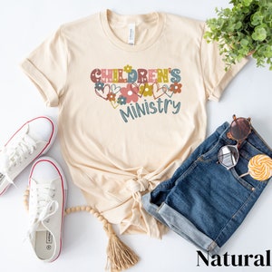 May include: A beige t-shirt with the text "Children's Ministry" in colorful letters with floral accents. The shirt is laid flat on a white surface with a pair of white sneakers, a pair of blue denim shorts, and a pair of sunglasses.