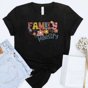 May include: Black t-shirt with the text "FAMILY MINISTRY" in colorful letters with flowers and hearts around the text.