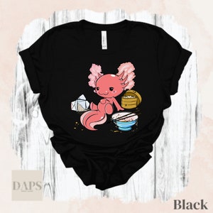 Peut inclure: T-shirt noir avec un axolotl rose en dessin anim&eacute; mangeant des nouilles dans un bol. L'axolotl est assis sur une table avec un r&eacute;cipient &agrave; emporter et des baguettes.