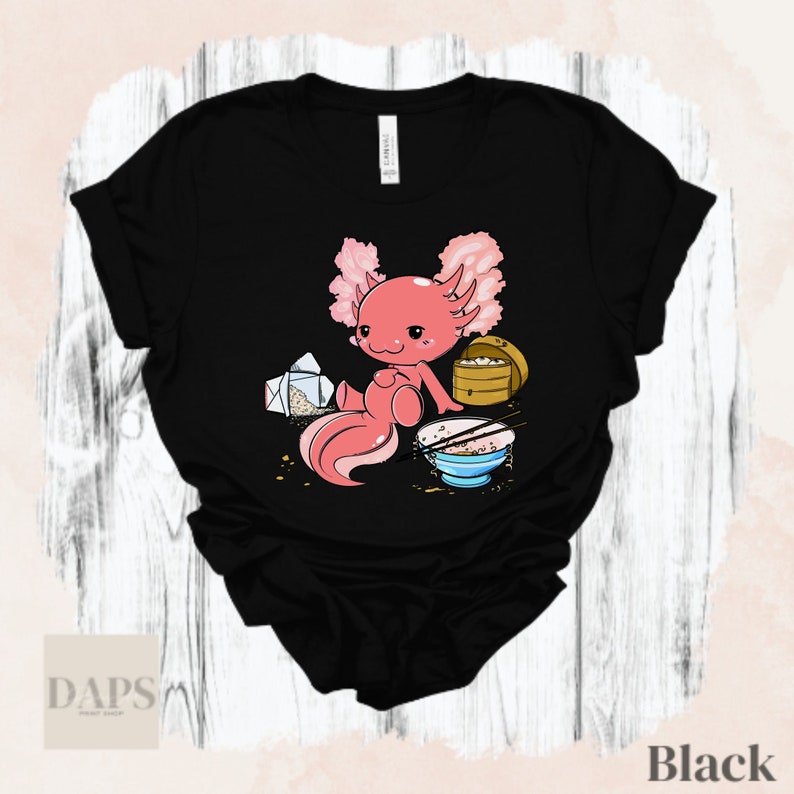 Peut inclure: T-shirt noir avec un axolotl rose en dessin anim&eacute; mangeant des nouilles dans un bol. L'axolotl est assis sur une table avec un r&eacute;cipient &agrave; emporter et des baguettes.
