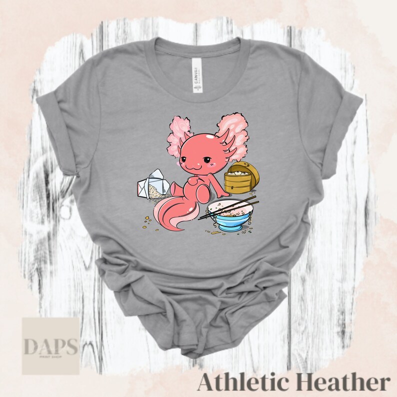 Peut inclure: Un t-shirt gris avec un axolotl de dessin anim&eacute; portant un bandeau rose et mangeant du riz dans un bol. L'axolotl est assis sur une surface blanche avec un r&eacute;cipient &agrave; emporter et des baguettes.