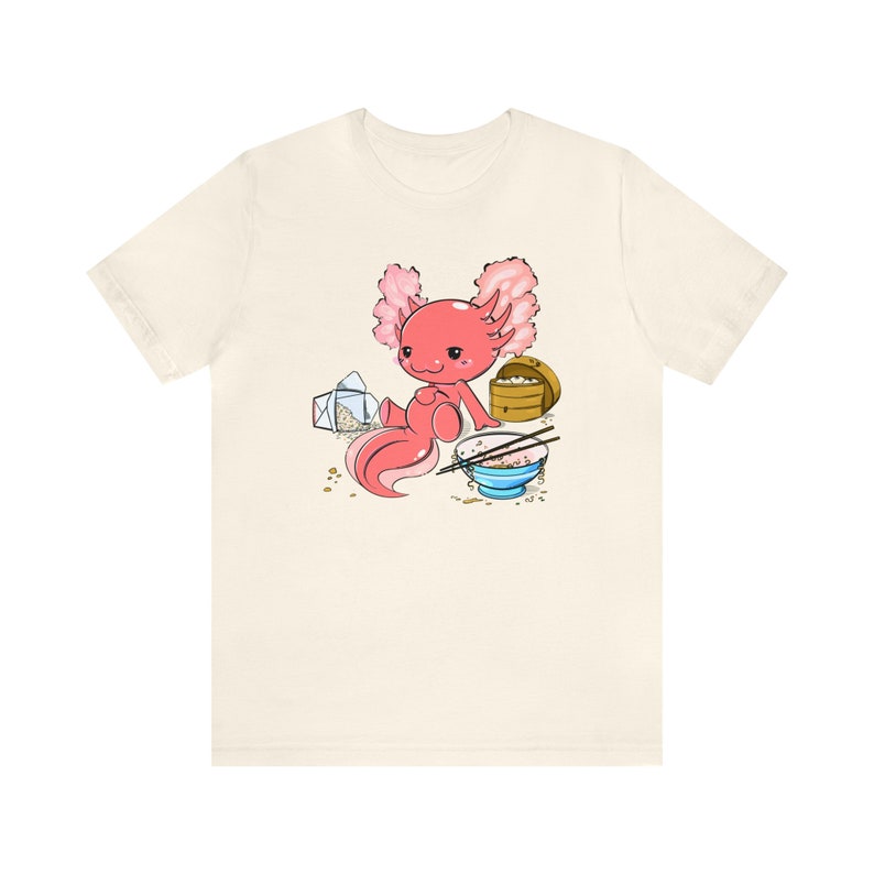Peut inclure: Un t-shirt blanc avec une illustration de dessin anim&eacute; d'un axolotl rose mangeant un bol de riz avec des baguettes. L'axolotl est assis sur un fond blanc avec une bo&icirc;te &agrave; emporter et une bo&icirc;te en bois.