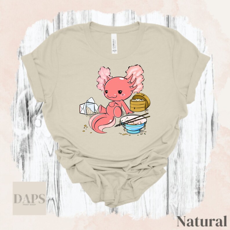 Peut inclure: Un t-shirt beige avec une illustration de dessin anim&eacute; d'un axolotl rose. L'axolotl est assis avec un bol de riz et des baguettes, une bo&icirc;te &agrave; emporter et une lettre.