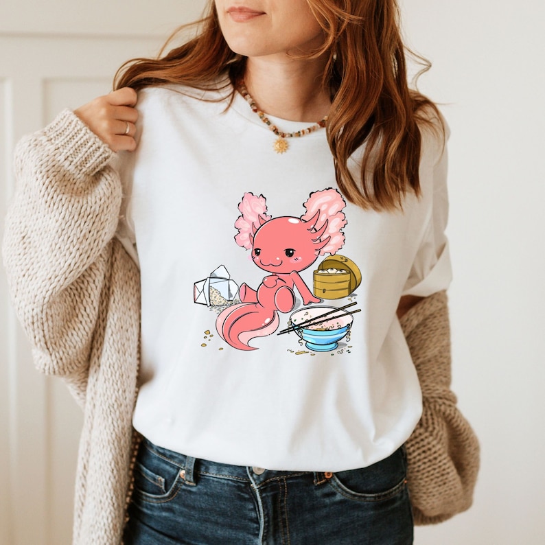 Peut inclure: T-shirt blanc avec une illustration de dessin anim&eacute; d'un axolotl rose portant un bol bleu avec des baguettes et du riz. L'axolotl est assis &agrave; c&ocirc;t&eacute; d'une petite bo&icirc;te en bois avec un couvercle et une bo&icirc;te &agrave; emporter blanche avec une enveloppe rose.