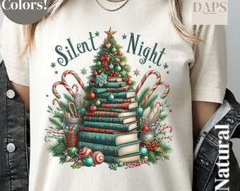 May include: Cream-colored t-shirt featuring a Christmas tree design made of stacked books, candy canes, and ornaments. The words "Silent Night" are written above the tree in a teal script. The shirt has a crew neck and short sleeves.