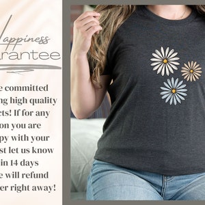 May include: A dark gray t-shirt with three white daisies with yellow centers. The daisies are arranged in a triangular pattern. The t-shirt has the text "Happiness Guarantee" printed on it.