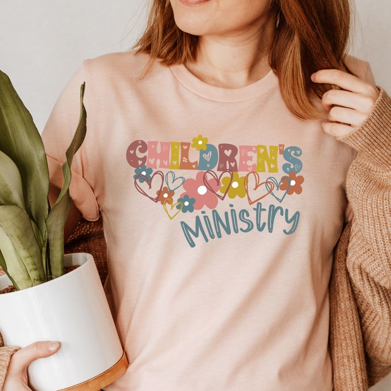 May include: A pink t-shirt with the text "CHILDREN'S MINISTRY" in colorful letters surrounded by hearts and flowers.