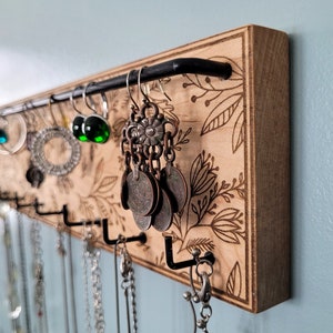 Wall mounted jewelry organizer made of solid maple wood and engraved with a floral pattern. There is a black steel bar for earrings to hang on and a series of hooks for necklaces and other accessories.