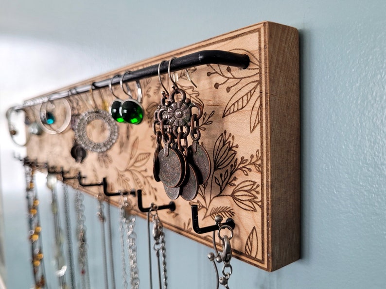 Wall mounted jewelry organizer made of solid maple wood and engraved with a floral pattern. There is a black steel bar for earrings to hang on and a series of hooks for necklaces and other accessories.
