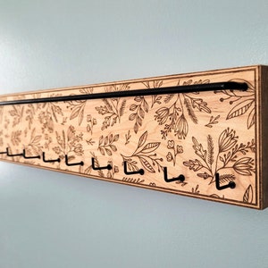 Wall mounted jewelry organizer made of solid maple wood and engraved with a floral pattern. There is a black steel bar for earrings to hang on and a series of hooks for necklaces and other accessories.