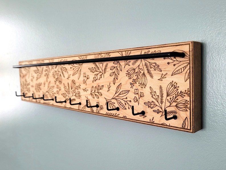Wall mounted jewelry organizer made of solid maple wood and engraved with a floral pattern. There is a black steel bar for earrings to hang on and a series of hooks for necklaces and other accessories.