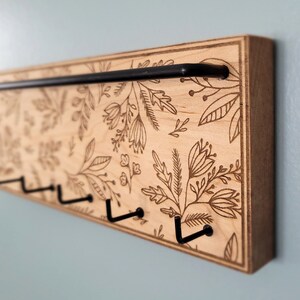 Wall mounted jewelry organizer made of solid maple wood and engraved with a floral pattern. There is a black steel bar for earrings to hang on and a series of hooks for necklaces and other accessories.