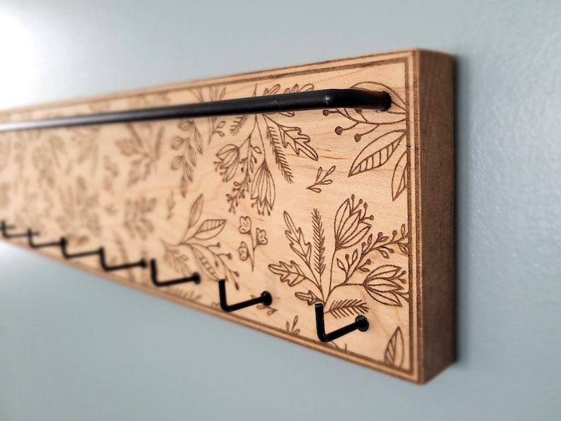 Wall mounted jewelry organizer made of solid maple wood and engraved with a floral pattern. There is a black steel bar for earrings to hang on and a series of hooks for necklaces and other accessories.
