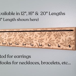 Wall mounted jewelry organizer made of solid maple wood and engraved with a floral pattern. There is a black steel bar for earrings to hang on and a series of hooks for necklaces and other accessories.