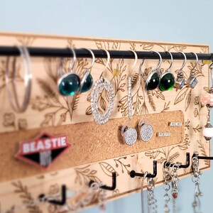 Wall mounted jewelry organizer made of solid maple wood and engraved with a floral pattern. There is a black steel bar for earrings to hang on, a cork insert for stud earrings and a series of hooks for necklaces and other accessories.