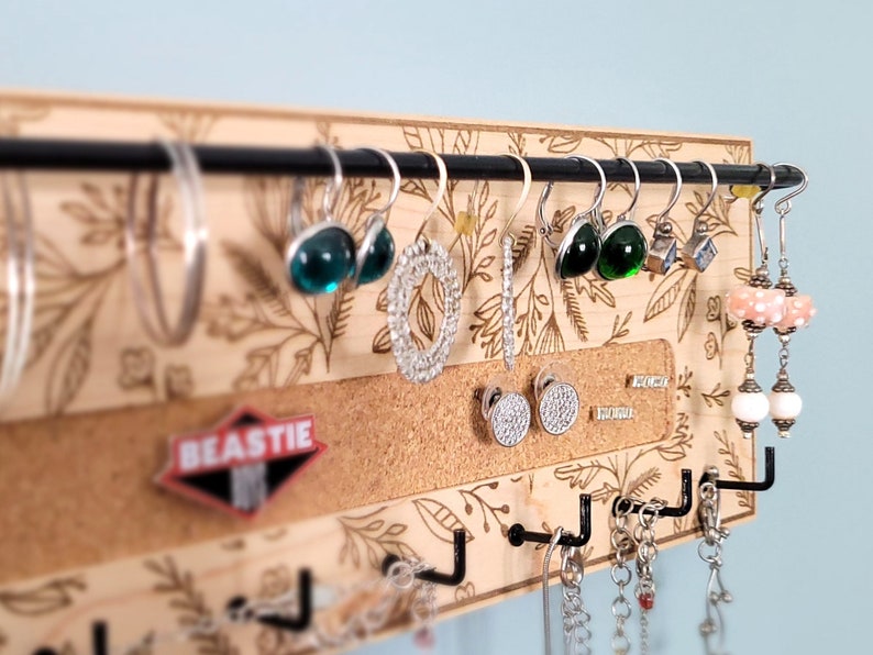 Wall mounted jewelry organizer made of solid maple wood and engraved with a floral pattern. There is a black steel bar for earrings to hang on, a cork insert for stud earrings and a series of hooks for necklaces and other accessories.