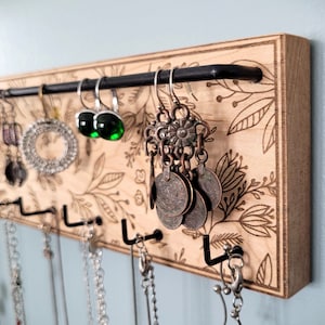 Wall mounted jewelry organizer made of solid maple wood and engraved with a floral pattern. There is a black steel bar for earrings to hang on and a series of hooks for necklaces and other accessories.