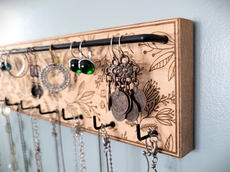 Wall mounted jewelry organizer made of solid maple wood and engraved with a floral pattern. There is a black steel bar for earrings to hang on and a series of hooks for necklaces and other accessories.