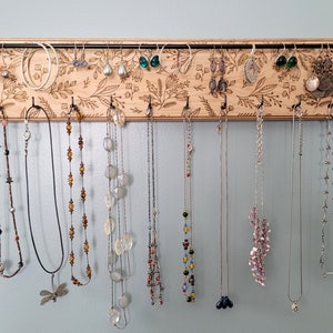 Wall mounted jewelry organizer made of solid maple wood and engraved with a floral pattern. There is a black steel bar for earrings to hang on and a series of hooks for necklaces and other accessories.