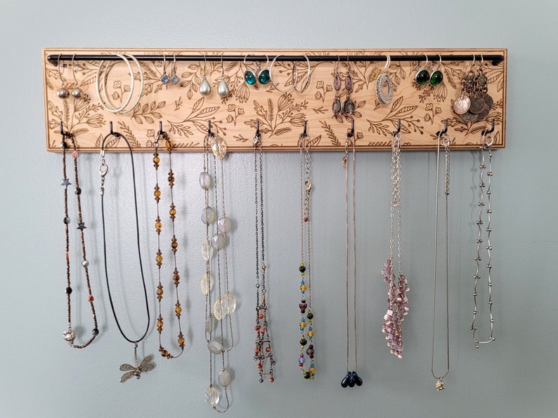 Wall mounted jewelry organizer made of solid maple wood and engraved with a floral pattern. There is a black steel bar for earrings to hang on and a series of hooks for necklaces and other accessories.