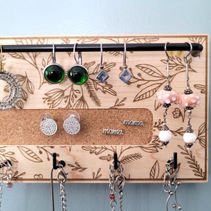 May include: A wooden jewelry organizer with hooks for necklaces and a corkboard section for earrings. The corkboard has a floral design and the words "mama" written on it. There are several pairs of earrings hanging from the hooks, including green, blue, and white earrings.