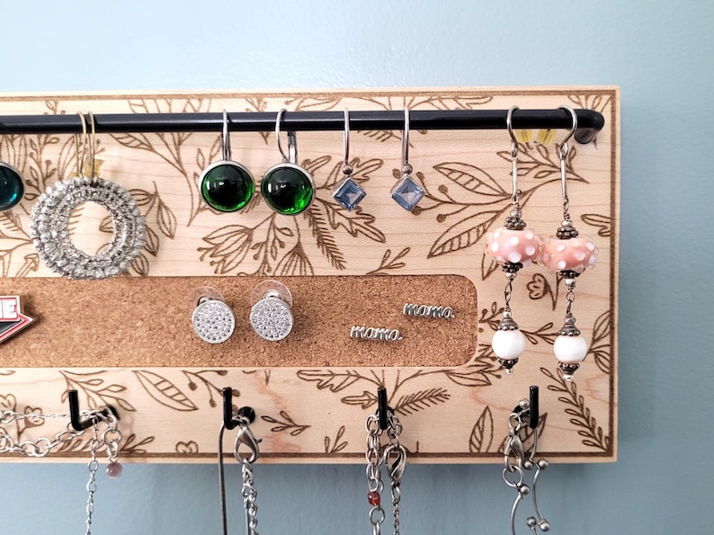 May include: A wooden jewelry organizer with hooks for necklaces and a corkboard section for earrings. The corkboard has a floral design and the words "mama" written on it. There are several pairs of earrings hanging from the hooks, including green, blue, and white earrings.