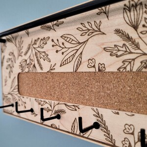 Wall mounted jewelry organizer made of solid maple wood and engraved with a floral pattern. There is a black steel bar for earrings to hang on, a cork insert for stud earrings and a series of hooks for necklaces and other accessories.