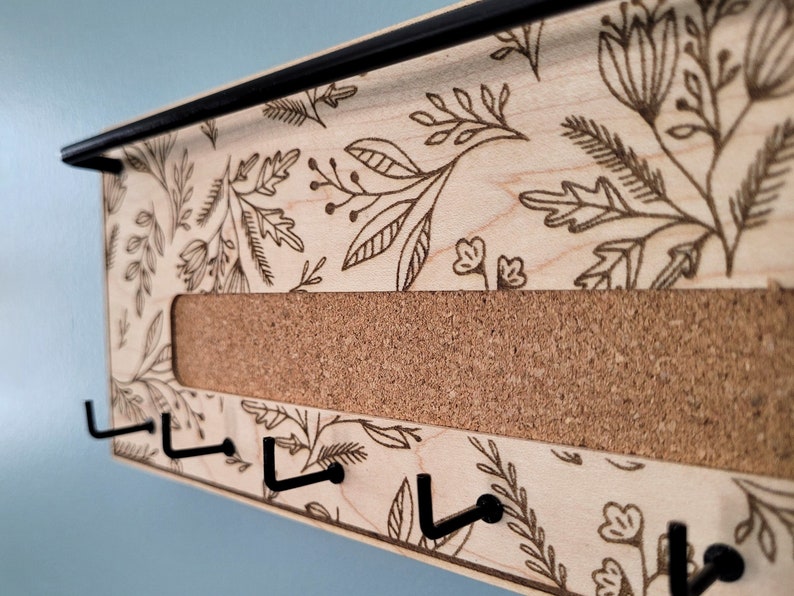 Wall mounted jewelry organizer made of solid maple wood and engraved with a floral pattern. There is a black steel bar for earrings to hang on, a cork insert for stud earrings and a series of hooks for necklaces and other accessories.