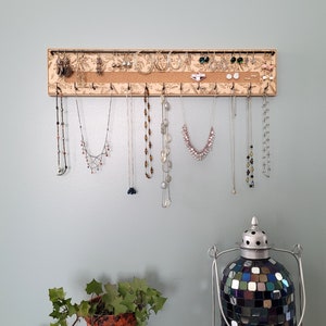 Wall mounted jewelry organizer made of solid maple wood and engraved with a floral pattern. There is a black steel bar for earrings to hang on, a cork insert for stud earrings and a series of hooks for necklaces and other accessories.