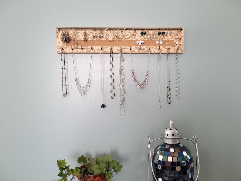 Wall mounted jewelry organizer made of solid maple wood and engraved with a floral pattern. There is a black steel bar for earrings to hang on, a cork insert for stud earrings and a series of hooks for necklaces and other accessories.