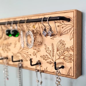 Wall mounted jewelry organizer made of solid maple wood and engraved with a floral pattern. There is a black steel bar for earrings to hang on and a series of hooks for necklaces and other accessories.