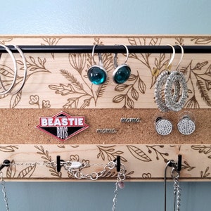Wall mounted jewelry organizer made of solid maple wood and engraved with a floral pattern. There is a black steel bar for earrings to hang on, a cork insert for stud earrings and a series of hooks for necklaces and other accessories.