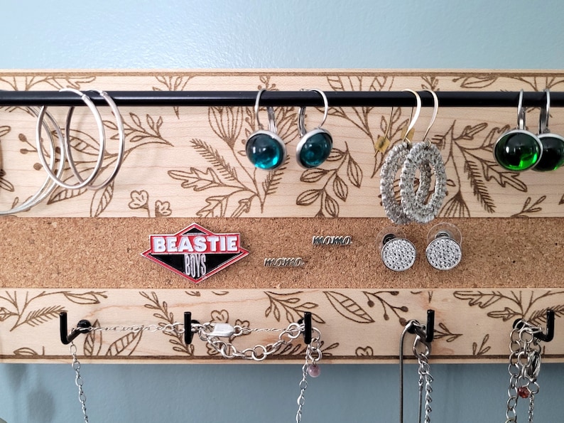 Wall mounted jewelry organizer made of solid maple wood and engraved with a floral pattern. There is a black steel bar for earrings to hang on, a cork insert for stud earrings and a series of hooks for necklaces and other accessories.