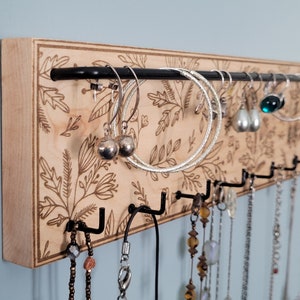 Wall mounted jewelry organizer made of solid maple wood and engraved with a floral pattern. There is a black steel bar for earrings to hang on and a series of hooks for necklaces and other accessories.