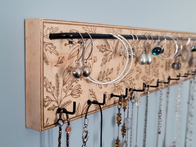 Wall mounted jewelry organizer made of solid maple wood and engraved with a floral pattern. There is a black steel bar for earrings to hang on and a series of hooks for necklaces and other accessories.