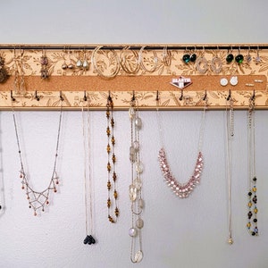 Wall mounted jewelry organizer made of solid maple wood and engraved with a floral pattern. There is a black steel bar for earrings to hang on, a cork insert for stud earrings and a series of hooks for necklaces and other accessories.