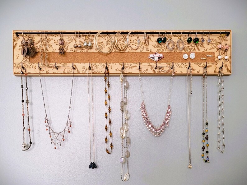 Wall mounted jewelry organizer made of solid maple wood and engraved with a floral pattern. There is a black steel bar for earrings to hang on, a cork insert for stud earrings and a series of hooks for necklaces and other accessories.