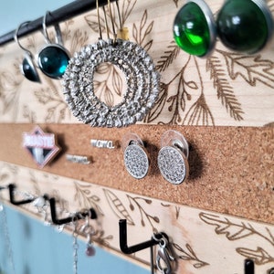 Wall mounted jewelry organizer made of solid maple wood and engraved with a floral pattern. There is a black steel bar for earrings to hang on, a cork insert for stud earrings and a series of hooks for necklaces and other accessories.
