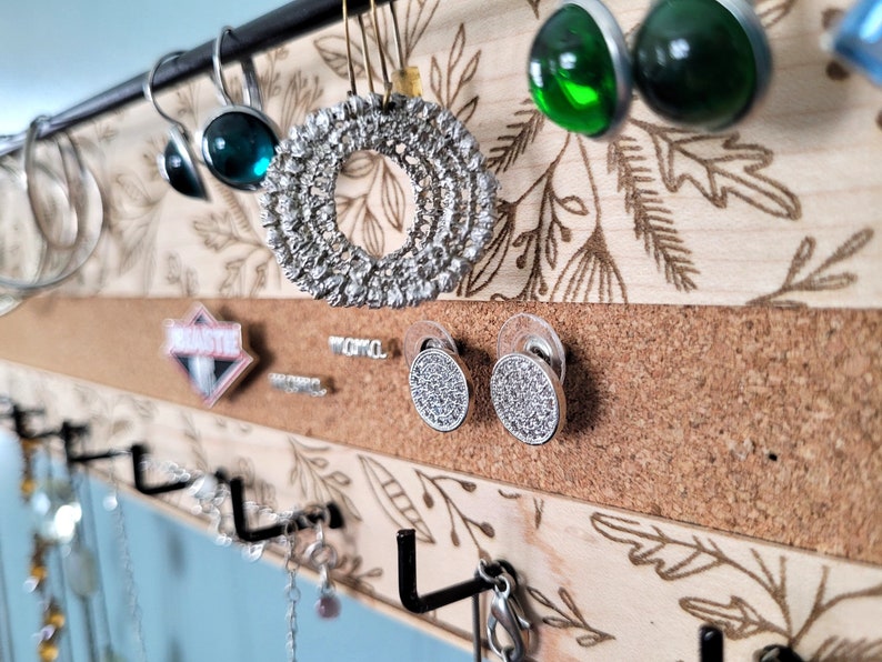 Wall mounted jewelry organizer made of solid maple wood and engraved with a floral pattern. There is a black steel bar for earrings to hang on, a cork insert for stud earrings and a series of hooks for necklaces and other accessories.