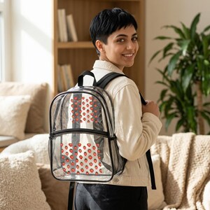 May include: Clear backpack with a black trim and straps. The backpack features a pattern of red strawberries on a white and black striped background. The backpack has a mesh pocket on the front and a zipper closure.