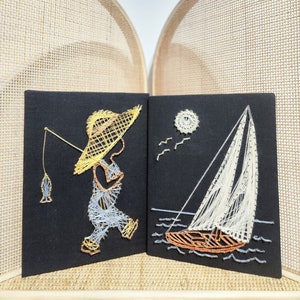 May include: Two framed string art pieces on a woven background. The left piece features a person in a straw hat fishing. The right piece features a sailboat with white sails and a brown hull.