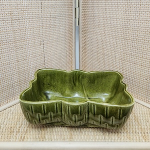 May include: A green ceramic leaf-shaped dish with a textured surface. The dish has a glossy finish and a divided interior. The exterior features a repeating pattern. This decorative dish is suitable for serving snacks or as a decorative accent.