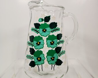 May include: A clear glass pitcher with a green and white floral design. The pitcher has a handle and a wide mouth.