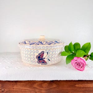 May include: A ceramic casserole dish with a lid, featuring a butterfly design. The dish is cream-colored with blue and red accents, and a floral pattern. A pink rose and green leaves are in the background.