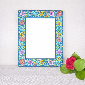 May include: A rectangular blue picture frame with a floral design in pink, yellow, and orange. The frame has a white interior and is decorated with colorful flowers and leaves. A red rose with green leaves is in the foreground.