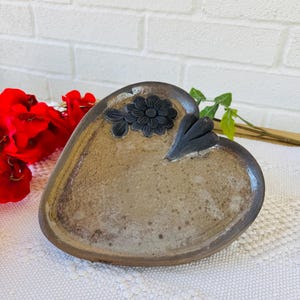 以下が含まれることがあります： 茶色の釉薬と黒い花柄が施されたハート型の陶器の皿。白いレースのランチョンマットの上に置かれています。