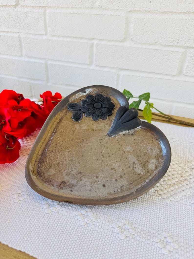 以下が含まれることがあります： 茶色の釉薬と黒い花柄が施されたハート型の陶器の皿。白いレースのランチョンマットの上に置かれています。