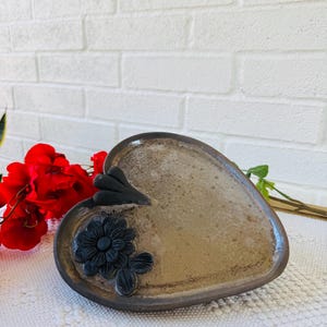 以下が含まれることがあります： 茶色の釉薬と黒い花柄のハート型のセラミック皿。手作りで素朴な風合い。