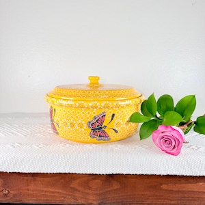 May include: A yellow ceramic casserole dish with a lid, decorated with a butterfly design. The dish has a dotted pattern and a handle on the lid. A pink rose and green leaves are in the background.